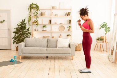 Kilo kaybı. Neşeli Afrikalı Amerikalı kız, evde tartıya çıkıp kendini tartıyor. Tam Uzunluk, Boş Alan