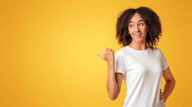 İyimser, iyi satış, teklif ya da terfi. Beyaz tişörtlü, gülümseyen Afro-Amerikan kıvırcık kadın harika bir şey görüyor, jestler, serbest alan, sarı arka planda izole edilmiş, stüdyo çekimi.