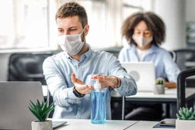 Ofisteki Coronavirus salgınında koruma. Koruyucu maskeli genç adam dizüstü bilgisayarda çalışıyor ve ellerini antiseptikle dezenfekte ediyor.
