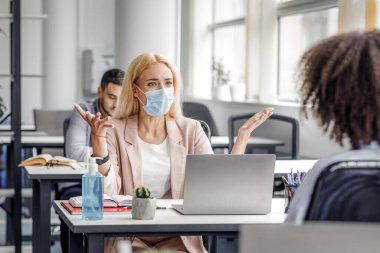 İş yerinde anlaşmazlık ve proje ile ilgili sorunlar. İş yerinde koruyucu maskeli kızgın bir kadın dizüstü bilgisayarla Afro-Amerikan bayana bağırıyor ve modern ofisin içinde hareketler yapıyor.