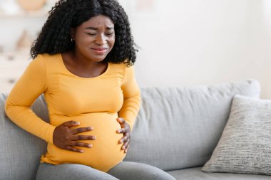 Black pregnant woman suffering from labor pains at home, copy space. African expecting lady having childbirth labor while sitting alone on sofa at home, child delivery concept