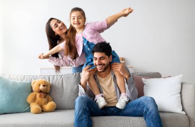 Neşeli, genç, doğulu ebeveynler oturma odasında çocukla eğleniyor, oynuyor, gülüyor ve birlikte vakit geçiriyorlar. Evde kızı olan mutlu bir Arap ailesi.