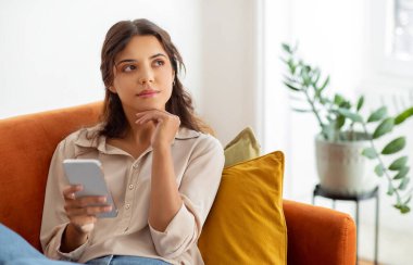 Akıllı telefonu tutan ve evdeki kanepede dinlenirken başka tarafa bakan düşünceli genç bir kadın. İç mekanda cep telefonu kullanan, düşünen ve bir şeyler planlayan güzel bir kadın.