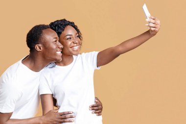 Neşeli Afro-Amerikan çift, sarı stüdyo arka planında akıllı telefondan selfie çekiyor. Mutlu genç siyah erkek ve kadın aşıklar bağlanırken, cep telefonu kullanarak fotoğraf çekiyorlar.