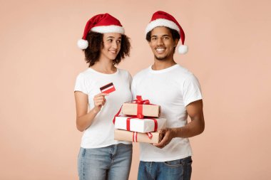 Noel için kolay ve hızlı hediyeler ve online alışveriş ile yeni yıl. Xmas şapkalı neşeli Afro-Amerikan çifti bir sürü hediye ve kredi kartı taşıyor, arka planda izole edilmiş, stüdyoda çekilmiş.