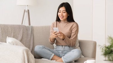 Free time. Asian girl checking social networks on phone, resting on sofa, panorama with free space