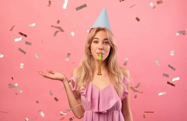 Doğum günü şapkalı güzel bir kadın pembe stüdyo arka planında konfeti düşüşüyle parti kornası çalıyor. Bayramları kutlayan bin yıllık olumlu bir bayan. Kutlama konsepti
