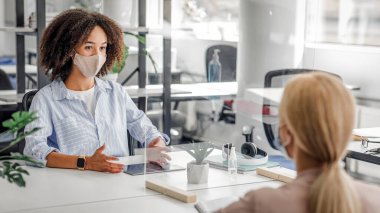Görüşme. Koruma maskesi takmış farklı etnik kökenlere sahip genç iş adamları COVID-19 enfeksiyonunu önlemek için bölme yoluyla birbirleriyle konuşuyorlar.
