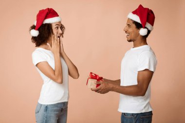 Evde kalıp yeni yılı ve Noel 'i birlikte kutlayalım. Gülümseyen Afrikalı Amerikalı Noel Baba şapkalı adam eşine hediye veriyor, hoş bir sürpriz, arka planda izole edilmiş, stüdyo çekimi