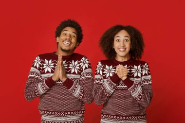 İyi şanslar ve yeni yıl ve Noel için büyük dilekler. Heyecanlı Afro-Amerikan çift, süveter içinde, gülümsüyor ve arzulamayı umuyor, dua ederken ellerini kavuşturuyor, kırmızı arka planda izole edilmiş, stüdyo çekimi