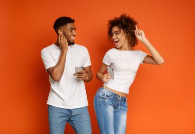 Neşeli bin yıllık siyah erkek ve kadın cep telefonları müzik dinleme ve her diğer, turuncu arka plana gülümseyen kulaklık