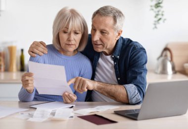 Serious couple of pensioners senior husband and wife sittting in front of computer at kitchen, reading letter, checking bills and correspondence, working on family budget together, copy space
