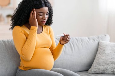 Hamile siyahi kadın baş ağrısı çekiyor, evde ateşi ölçüyor. Hasta bir Afrikalı Amerikalı kadının şakağına dokunmasını ve elindeki termometreye bakmasını bekliyor.