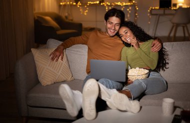 Genç eşler dizüstü bilgisayarda film izleyip patlamış mısır yiyorlar. Akşam evde koltukta dinleniyorlar. Lolow Light. Mutlu Çift Hafta Sonu Gecesinde Çevrimiçi Filmden Hoşlanıyor