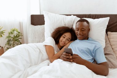Rahatlayın ve evde birlikte dinlenin yatakta, hafta sonlarında ve boş zamanlarınızda. Neşeli, gülümseyen, Afro-Amerikan çift sabah rahat yatak odasında sarılıp, akıllı telefonlara bakıyorlar.