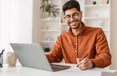 Gözlüklü gülümseyen genç bir Arap 'ın portresi dizüstü bilgisayar kullanıyor, masada oturuyor, deftere yazıyor. Uzaktan çalışan, internetten Webinar eğitimi alan, belge imzalayan mutlu bir adam.