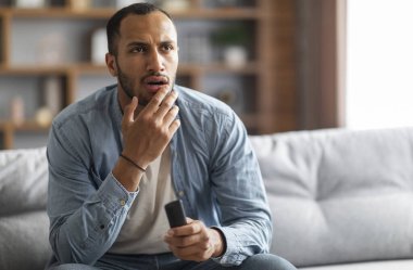 Shocked Black Man With Remote Controller In Hand Sitting On Couch At Home, Confused Young African American Guy Watching Tv Show In Living Room, Emotionally Reacting To Shocking Content, Copy Space