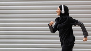 Mütevazı spor kıyafetleri ve kablosuz kulaklıkları olan fit Müslüman kadın dışarıda spor yapan tesettürlü atletik İslamcı kadın koşucunun yan görüntüsü, fotokopi alanı olan panorama.
