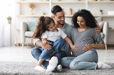 Mutlu Ortadoğu aile babası, hamile anne ve anaokulu çocuğu birlikte poz veriyor, evde oturuyor, bebeği bekliyor, küçük tatlı bir kız hamile annesine dokunuyor, büyük göbeğine, kopyalama alanına.