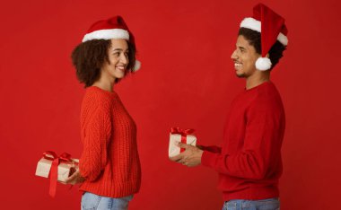 Birbirine aşık çift yeni yıl hediyelerini değiş tokuş ediyor. Binlerce yıllık Afro-Amerikan erkeği ve Noel Baba şapkalı kadın birbirlerine bakıyorlar ve kırmızı arka planda izole edilmiş kutuları tutuyorlar.