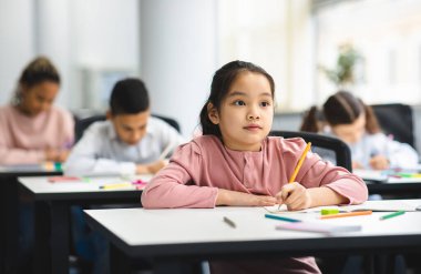İlkokul. Gülümseyen, notlar alan ve egzersiz defterinde yazan şirin Asyalı kız portresi. Yeni Bir Şey Öğrenen Farklı Çocuklar Grubu ile Küçük Sınıflar Sınıfı