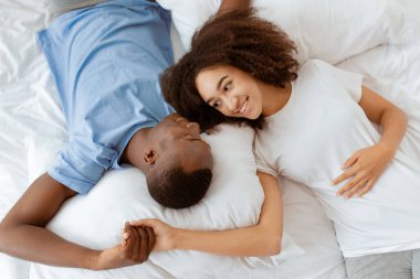 Evde birlikte mutlu olmak, hafta sonları dinlenmek ve rahatlamak. Gülümseyen bin yıllık Afro-Amerikan kadın ve erkek yumuşak beyaz yatağa uzanıp birbirlerine bakıyorlar yatak odasında, üst manzarada, boş yerde.