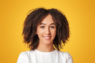 Modelin güzel tatlı gülümsemesi. Bin yıllık gülümseyen Afro-Amerikan kadın portresi beyaz tişörtlü kıvırcık saçlı, kameraya bakan, sarı arka planda izole edilmiş, stüdyo çekimi, serbest alan.