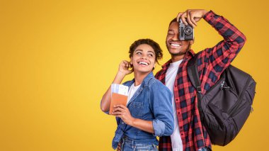 Seyahat et. Fotoğraf makineli neşeli Afrikalı turistler ve sarı stüdyo arka planında tatil pozu için hazırlar.