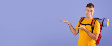 Kamp malzemeleriyle gülümseyen milenyum adamının portresi mavi stüdyo arka planında boş bir alanı işaret ediyor. Havalı genç adam seyahat, turizm ve panorama reklamları yapıyor.