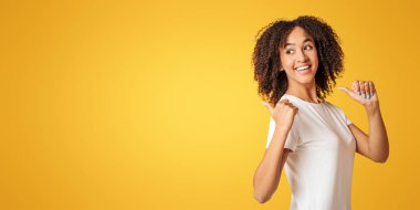 Öğrenci, blogcu harika bir teklif sunuyor. Beyaz tişörtlü kıvırcık saçlı, sarı arka planda izole edilmiş başparmaklarını boş alana gösteren binlerce yıllık mutlu Afro-Amerikan kadını.