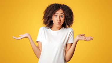 Yüz ifadeleri ve insani duygular. Beyaz tişörtlü, kıvırcık saçlı, kafası karışmış genç Afro-Amerikan kadın, sarı arka planda izole bir şekilde kollarını yana açıyor.
