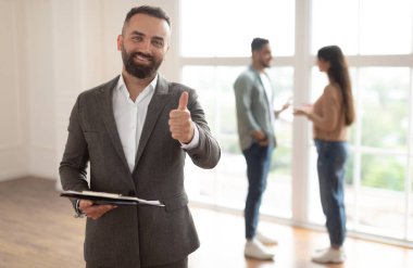 Başarılı bir anlaşma, Ev Sahipliği Konsepti. Gülümseyen profesyonel emlakçının portresi Başparmağı işaret gibi gösteren, seçici odaklanma. Bulanık Arkaplanda Konuşan Alıcılar Çifti