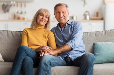 Yaşlı eşlerin el ele tutuşması, koltukta oturup kameraya gülümseyişi ve evde zaman geçirmesi. Ön Manzara. Mutlu Evlilikler ve Yaşam Tarzı