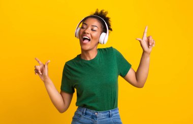 Vücudunu oynat. Gülümseyen afro kızın portresi kulaklıkla müzik dinleyen, gözleri kapalı dans eden ve işaret eden