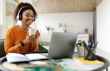 Laptop kullanan, elinde fincan tutan ve kahve içen gülümseyen siyah bir kadının portresi. Kablosuz kulaklıkla gülen kadın çay masasında oturuyor, video, film ya da webinar izliyor, ara veriyor.