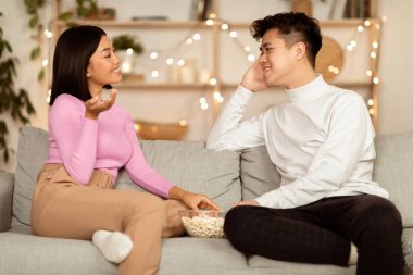 Neşeli Asyalı çift içeride koltukta oturmuş flört ediyor ve eğleniyor ve eğleniyorlar. Hafta sonu Akşam Yemeği, Flört ve Romantik İlişki Konsepti.