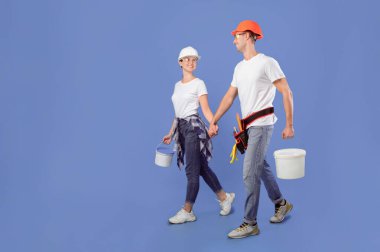 Güvenlik gözlüklü ve şapkalı genç eşler mavi arka planda boya malzemeleriyle yürüyorlar, milenyum işçi çifti apartman onarımı ve ev yenilemesi için hazır, tam boy çekim, fotokopi alanı