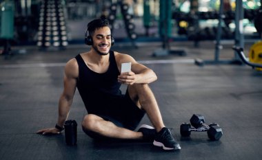 Fitness Uygulaması. Spor salonunda antrenmandan sonra dinlenirken Smartphone kullanan gülümseyen Orta Doğulu sporcu, Yakışıklı Arap Atletizm Eğitimli Yeni Mobil Uygulamaları Gözlemliyor, Boşluğu Kopyala