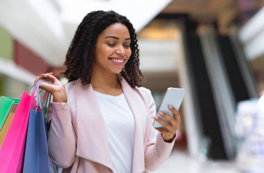 İndirimli uygulama. Gülümseyen Afrikalı Amerikalı Kadın alışveriş merkezinde akıllı telefon kullanıyor, indirimleri kontrol ediyor, bedava yer açıyor.