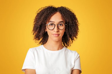 Çalışmaya hazır inek öğrenci. Beyaz tişörtlü, gözlüklü, kıvırcık saçlı, sarı arka planda, stüdyo çekimlerinde izole edilmiş beyaz saçlı Afro-Amerikan kadını.