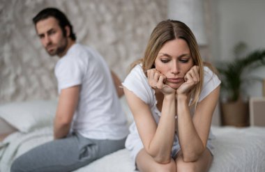 Cinsel sorunlar, ilişki krizi konsepti. Stresli genç kadın yatakta oturuyor, aile sorunları yüzünden bunalmış, arka planda gücenmiş kocası, boş bir alan.