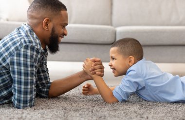 Mutlu siyah baba ve oğlunun evde eğlenirken yerde yatıp bilek güreşi yapmasının yan görüntüsü. Küçük çocuk bilek güreşi yapıyor. Gerçek Bir Erkek Kavramı Büyütmek.