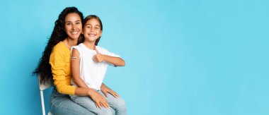 Çocuk Aşısı. Neşeli Orta Doğulu Anne ve Kızı, Aşılanmış Kol ve Mavi Arkaplan 'da Covid-19' a karşı aşıyı onaylayan el kol hareketi gibi. Panorama, Boşluk Boşluğu