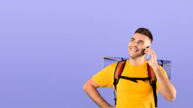 Turist vitesli çekici milenyum çocuğu mavi stüdyo arka planında akıllı telefondan konuşuyor. Mutlu genç adam seyahatleri sırasında cep telefonuyla dostça sohbet ediyor.