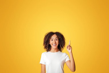 Harika bir fikir ya da iyi bir teklif. Gülümseyen, kıvırcık saçlı, beyaz tişörtlü, heyecanlı Afro-Amerikalı kadın boş bir yeri işaret ediyor, sarı arka planda izole edilmiş, stüdyo çekimi, kesilmiş.