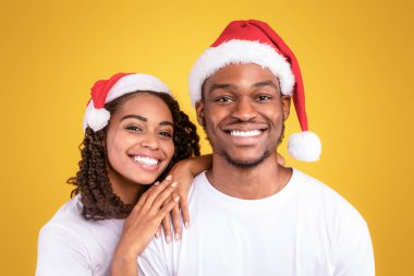 Noel Baba Şapkalı Neşeli Siyah Çift 'in Noel Portresi Sarı Stüdyo Arkaplanda Poz Veriyor, Kameraya Gülümsüyor. Xmas, Yeni Yıl ve Kış Tatilleri Kutlaması. Panorama