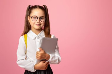 Okul başvuruları. Gözlüklü ve üniformalı arkadaş canlısı kız tablet tutuyor, pembe arka planda izole edilmiş, stüdyo çekimlerinde.