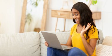 Çevrimiçi iletişim kavramı. Evdeki kanepede oturan neşeli genç siyahi kadın dizüstü bilgisayar başında ailesi, arkadaşları ya da iş arkadaşlarıyla konuşuyor. Güzel Afrikalı Amerikalı kadın internette sohbet ediyor.