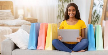 Ticaret konsepti. Alışveriş torbaları taşıyan Milenyum siyahi kadın çevrimiçi, kapalı mekanlarda ürünler almak için dizüstü bilgisayar kullanıyor. Güzel Afrikalı Amerikalı kadın internetten bir şeyler alıyor.