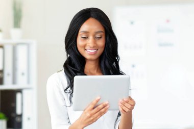 Uzaktan çalışma konsepti. Tablet bilgisayar kullanan neşeli siyahi kadın, modern ofisteki internette bilgi arıyor. Afrikalı Amerikalı girişimci çevrimiçi iş görüşmeleri yapıyor, çalışanlarla konferans yapıyor.